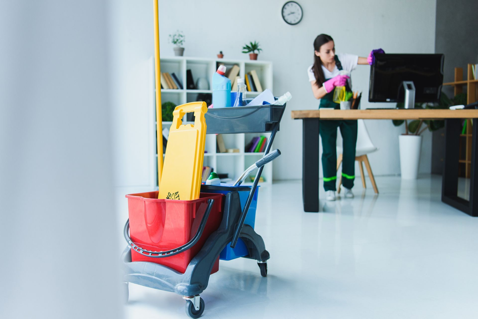 Office cleaning cart with supplies and worker disinfecting desk in modern workspace interior setting.