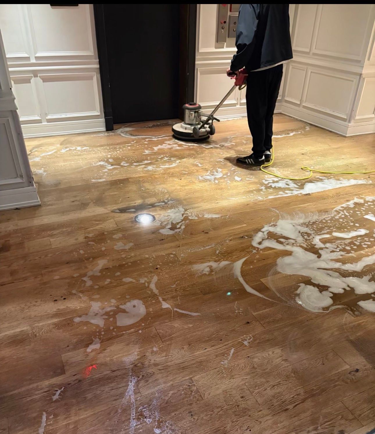 Person operating a floor buffer on a varnished wood floor in a paneled hallway.