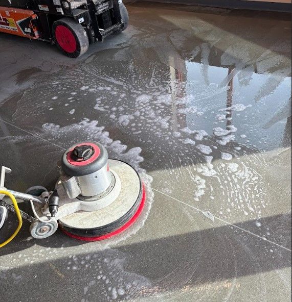 Floor being cleaned with a rotary scrubber; suds and water on a dark stone surface, machine in upper left.