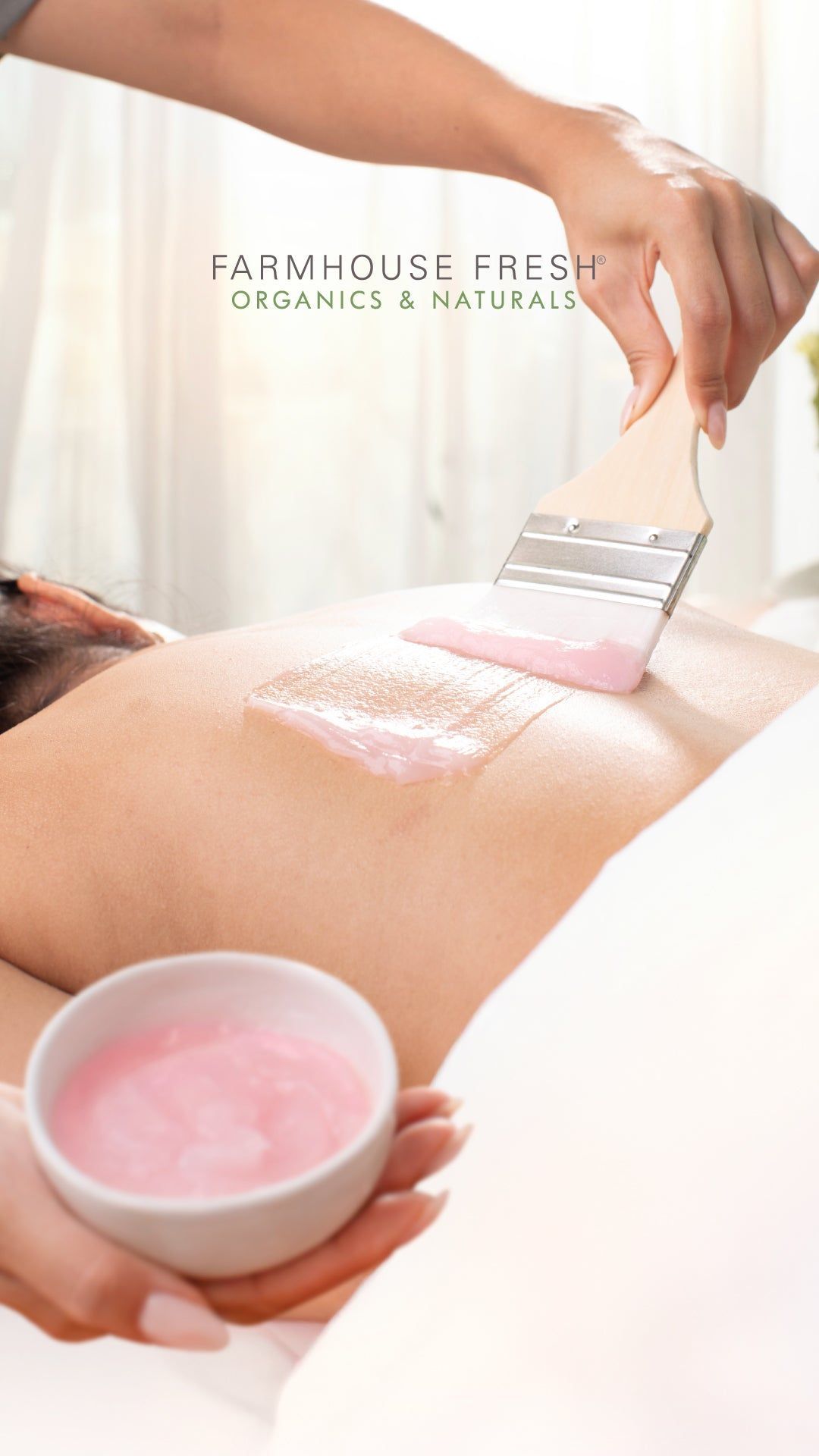 A person uses a brush to apply a pink, creamy treatment onto someone's back from a small bowl.