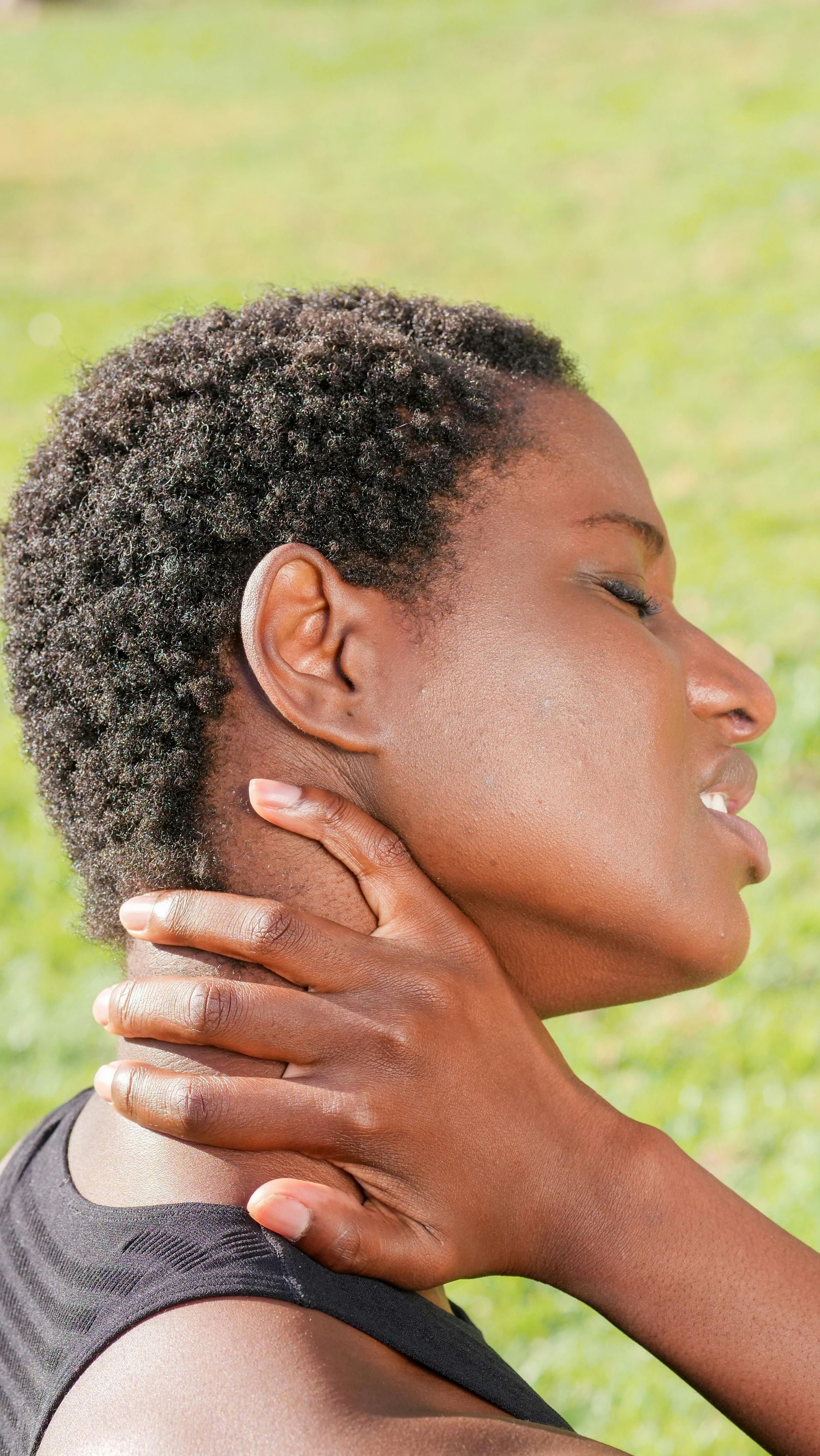 A person wearing a black shirt outdoors holds the back of their neck with their hand, appearing to be in pain or discomfort.