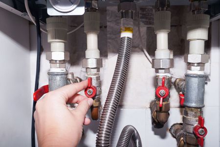 Hand turns red valve on under-sink plumbing with pipes and flexible hose.