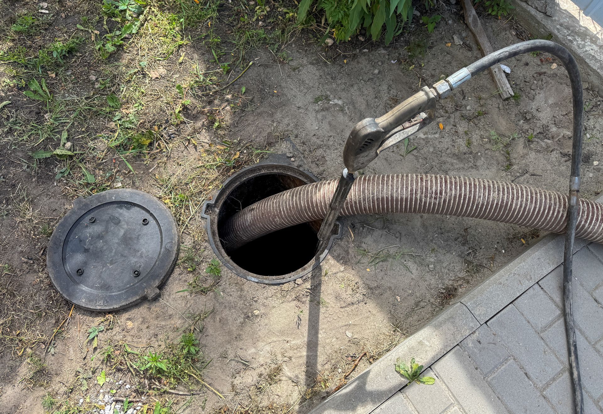 Open manhole with a hose inserted beside a sidewalk and round cover on the ground