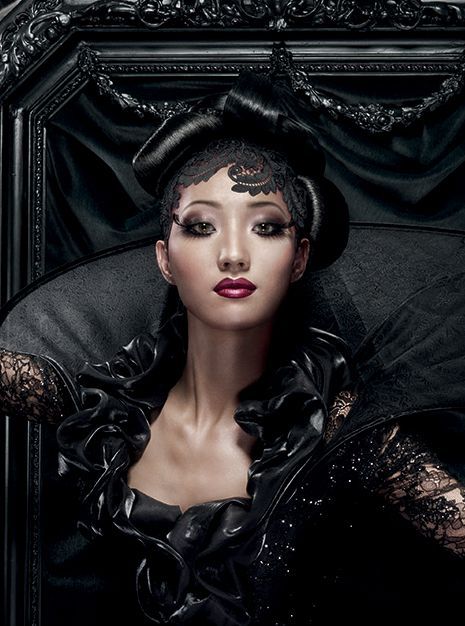 Vrouw in een uitgebreide zwarte jurk, met make-up en kapsel, omlijst door een sierlijke zwarte achtergrond.
