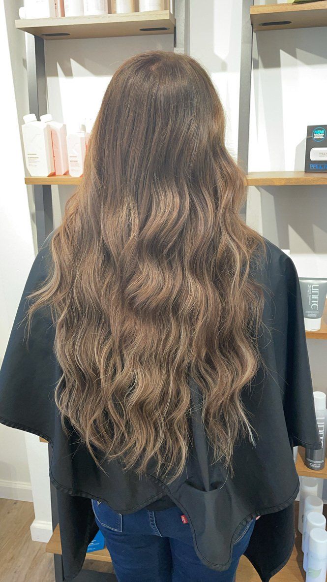 A woman with long brown hair is standing in a salon wearing a black cape.