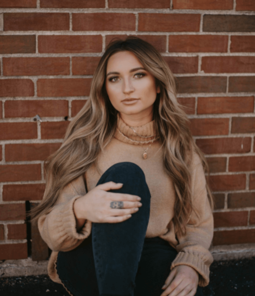 A woman in a tan sweater is sitting in front of a brick wall