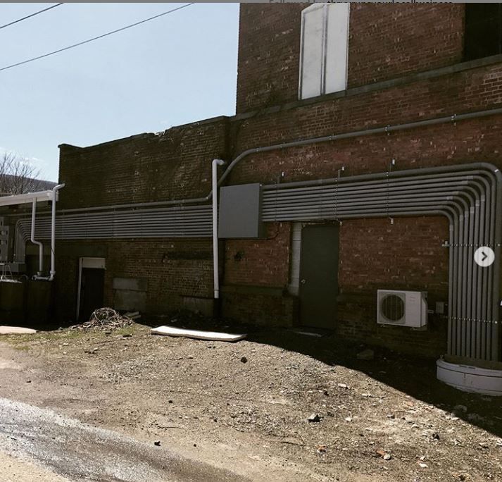 Brick building exterior with exposed utility conduits and an air conditioner unit along a dirt lot