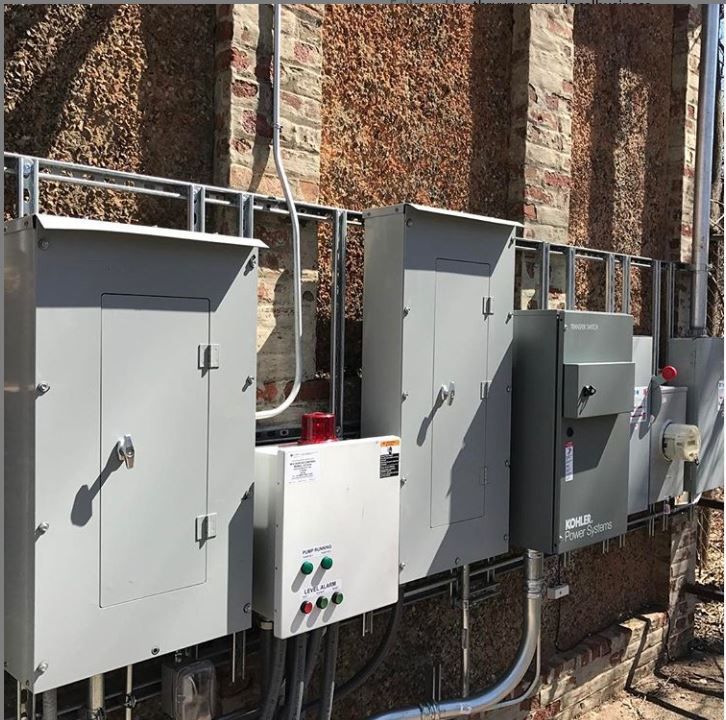 Row of gray electrical control panels and meters mounted on a brick wall outdoors.
