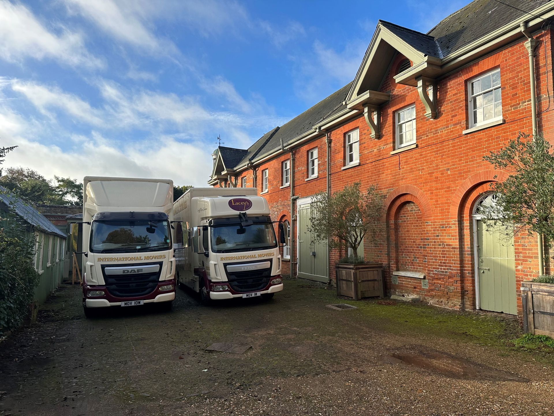 Two white and burgundy box trucks parked in front of a brick building with arched doorways.