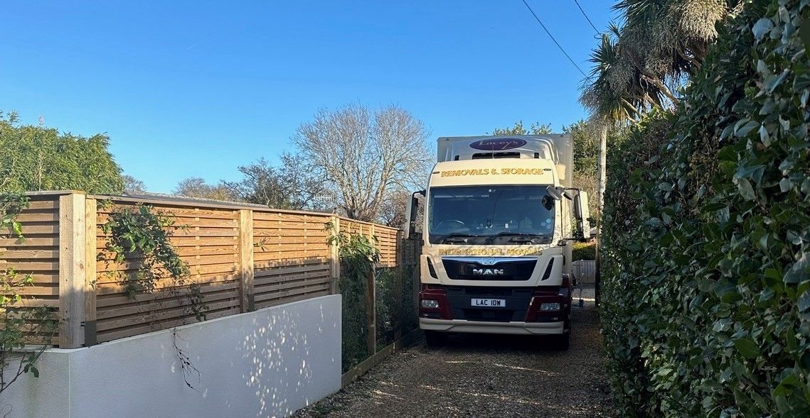 A truck is parked in a driveway. Wooden fence on the left, bushes on the right. Blue sky in the background.