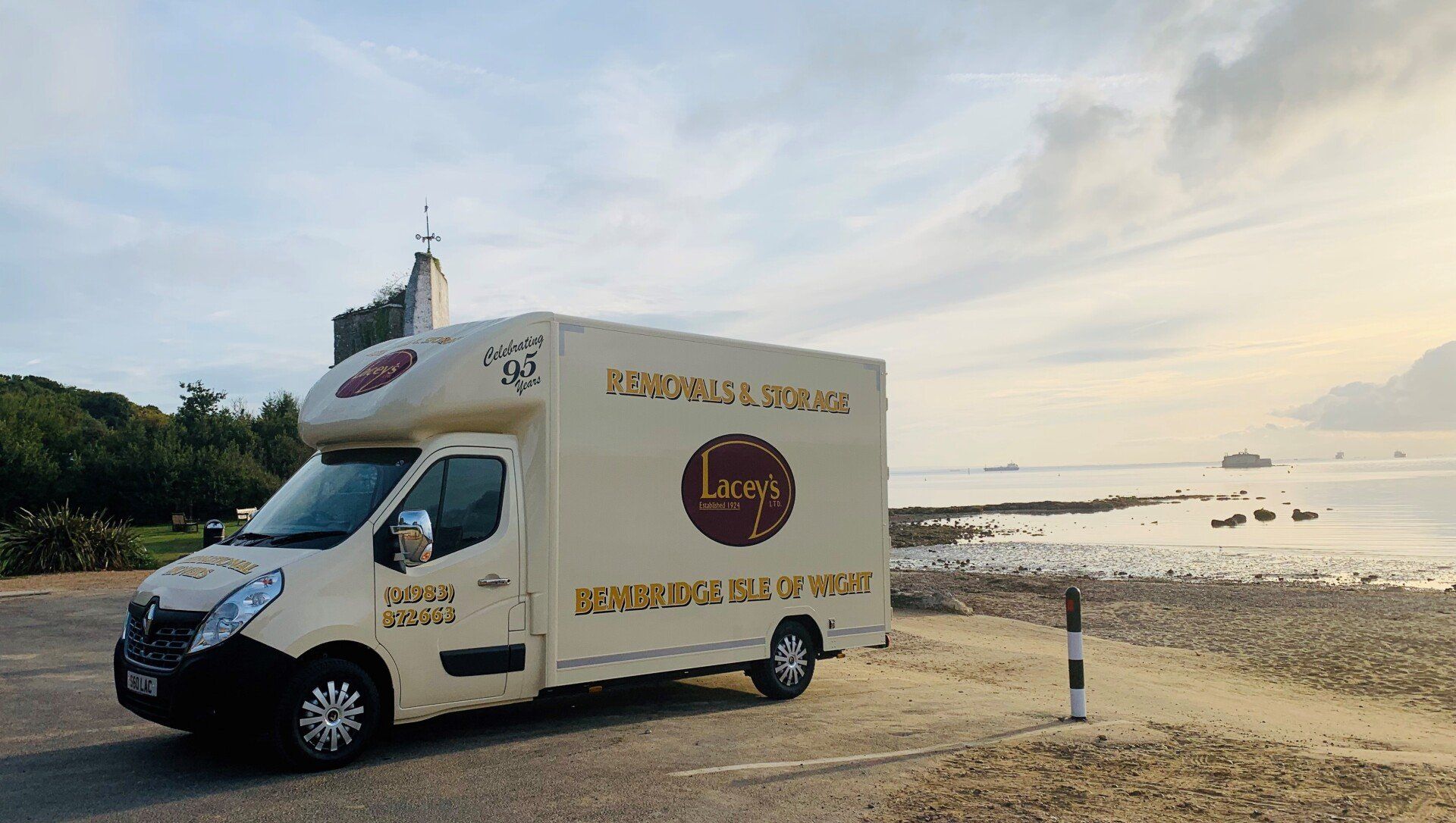Cream-colored food truck parked by a rocky beach. The ocean and a distant building are visible under a cloudy sky.