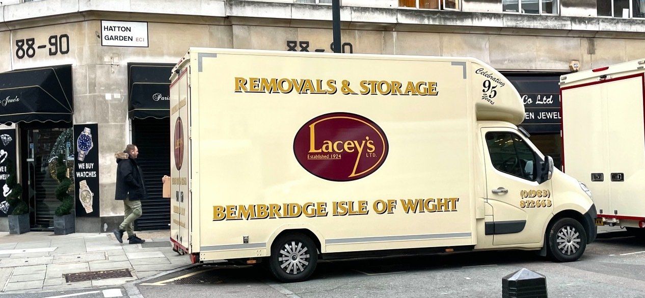 A man carrying a box walks from a cream-colored Lacey's removal van toward a building.