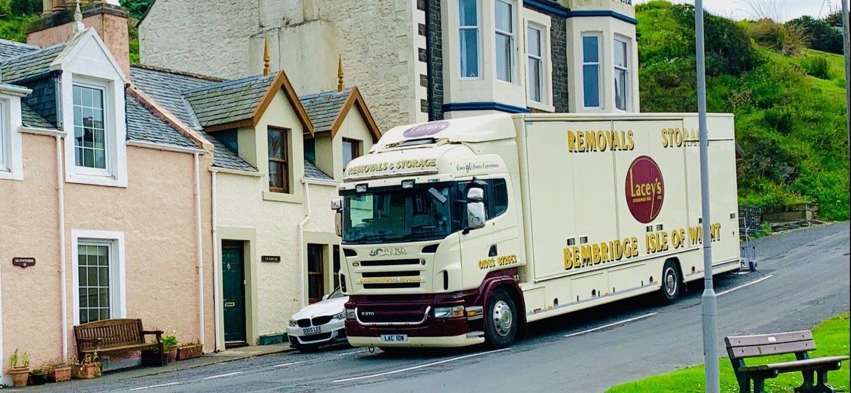 Moving truck parked on a street with small buildings and a grassy hill in the background.