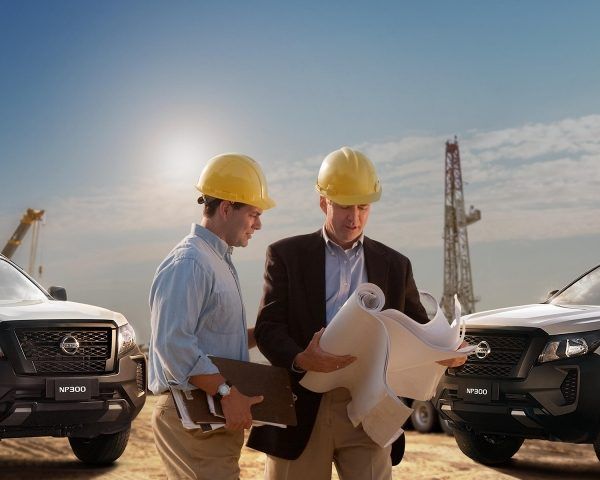 Dos ingenieros con cascos de seguridad revisan planos entre camionetas en una obra en construcción.