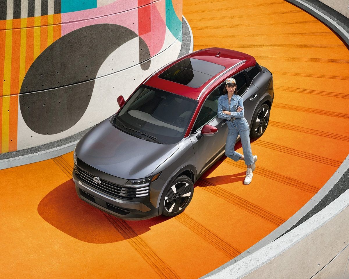 Persona posando junto a un SUV rojo y negro en una rampa en espiral naranja con un colorido mural de fondo.