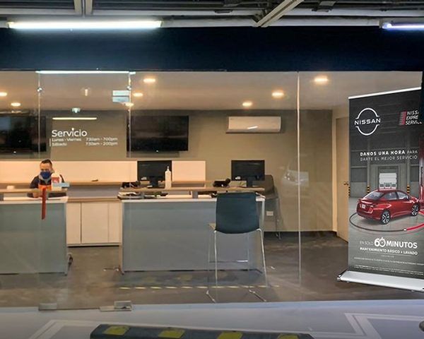 Mostrador de servicio de oficina moderno con mampara de vidrio, escritorios, sillas y una persona sentada a la izquierda.