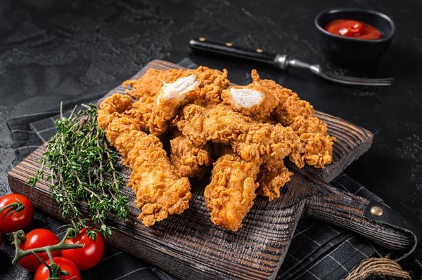 Fried chicken strips on a wooden cutting board with tomatoes and ketchup.