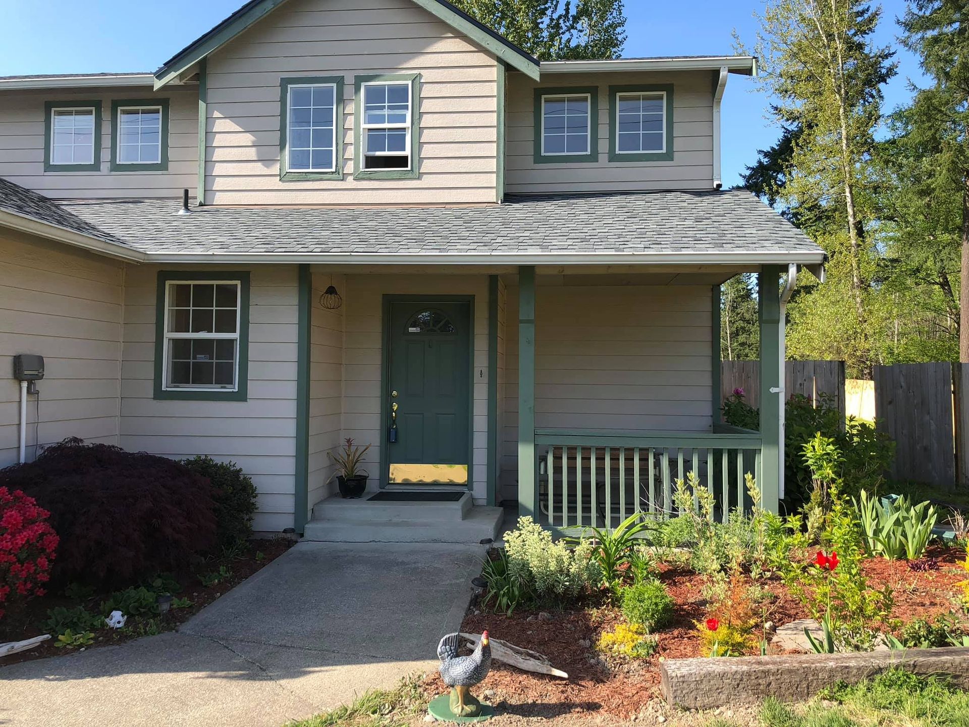 Two-story beige house with a front porch, green door, and colorful flower garden in front