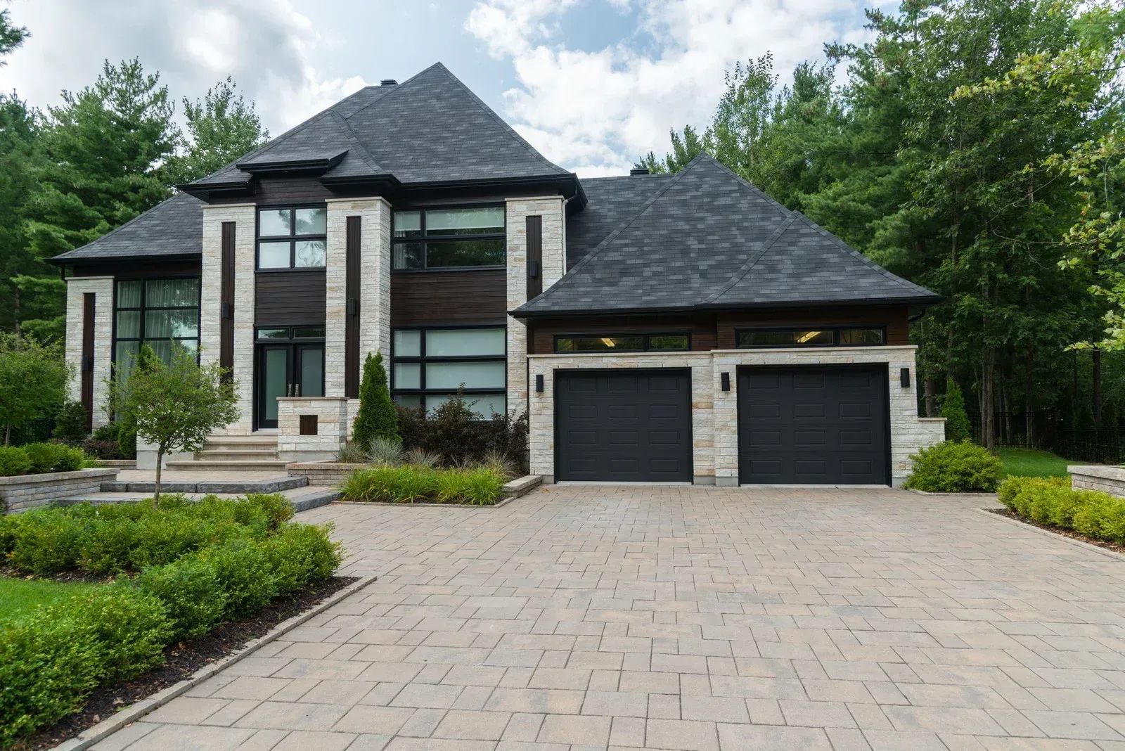 Modern two-story house with stone facade, dark roof, and double garage, viewed from the driveway