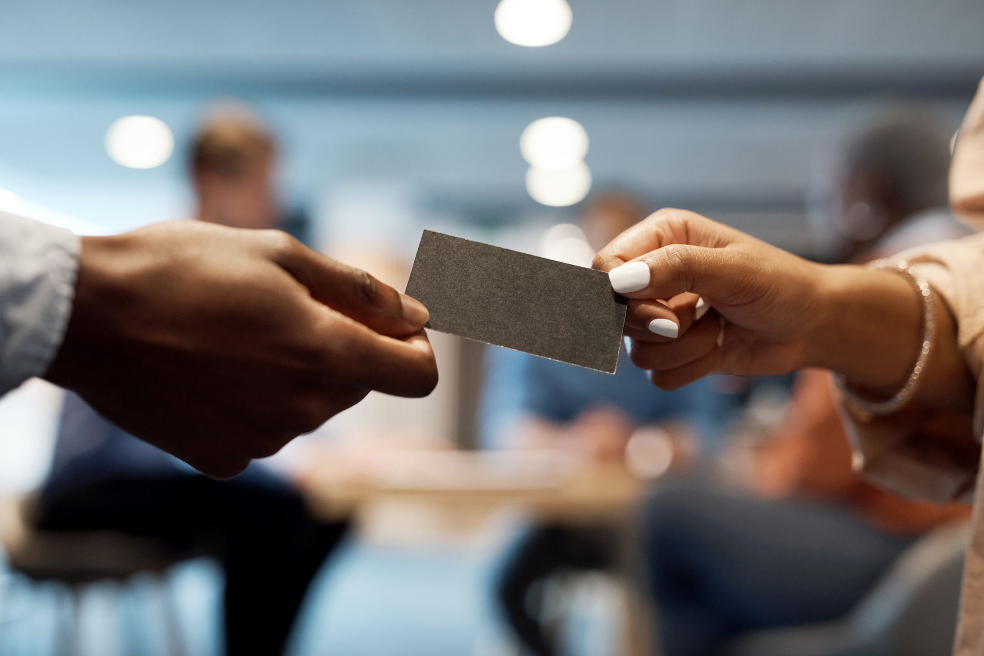 Two hands exchanging a dark business card. Other people blurred in the background, indoors.