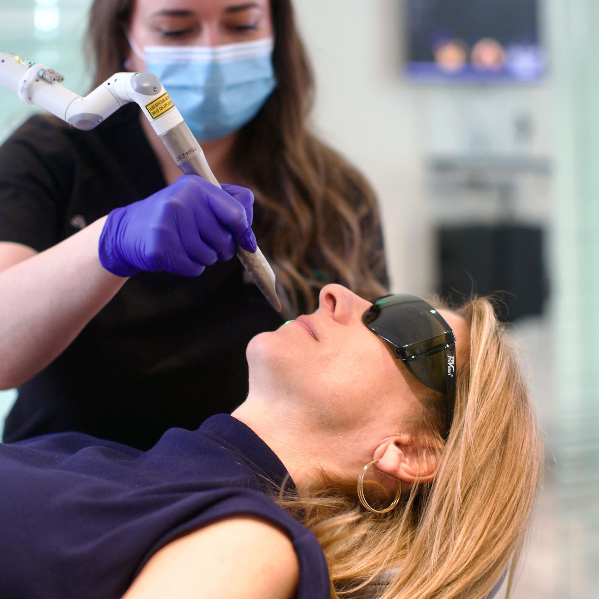 A medical professional in a mask and gloves uses a laser device on a woman's face, who wears protective eyewear in a clinic setting.