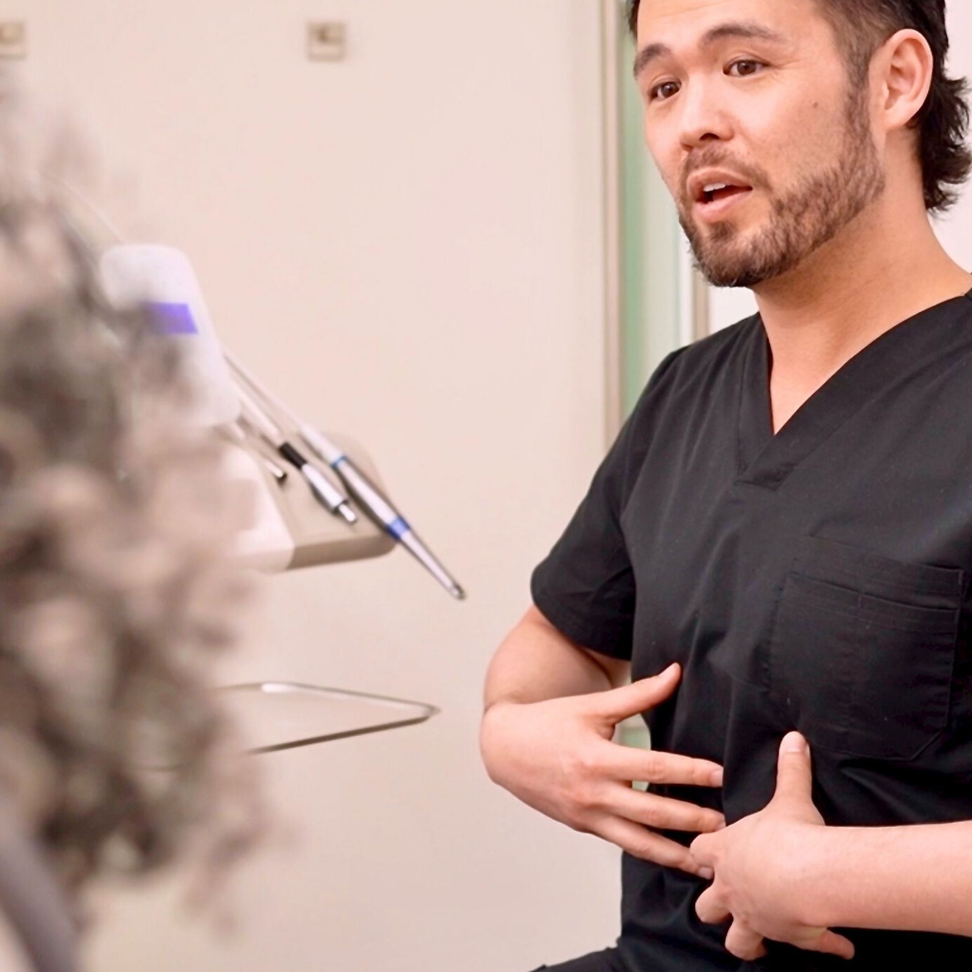 Man in black scrubs gestures toward his abdomen in a medical setting, near dental equipment. He has dark hair, a beard, and is speaking.