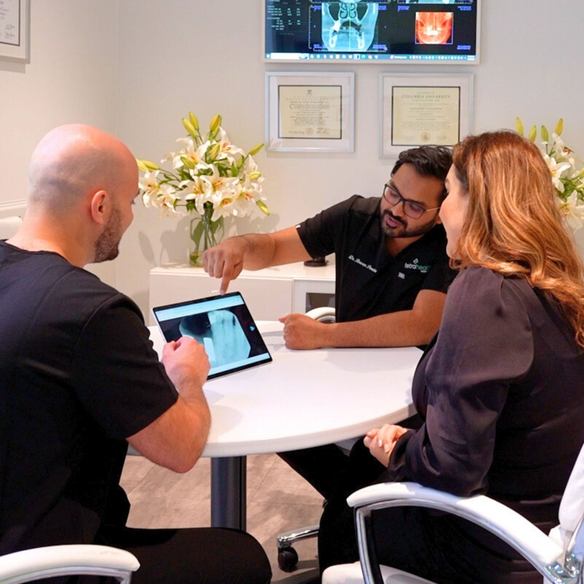 Three people in a dental office reviewing an X-ray on a tablet. A doctor points to the screen, explaining the results to a patient.