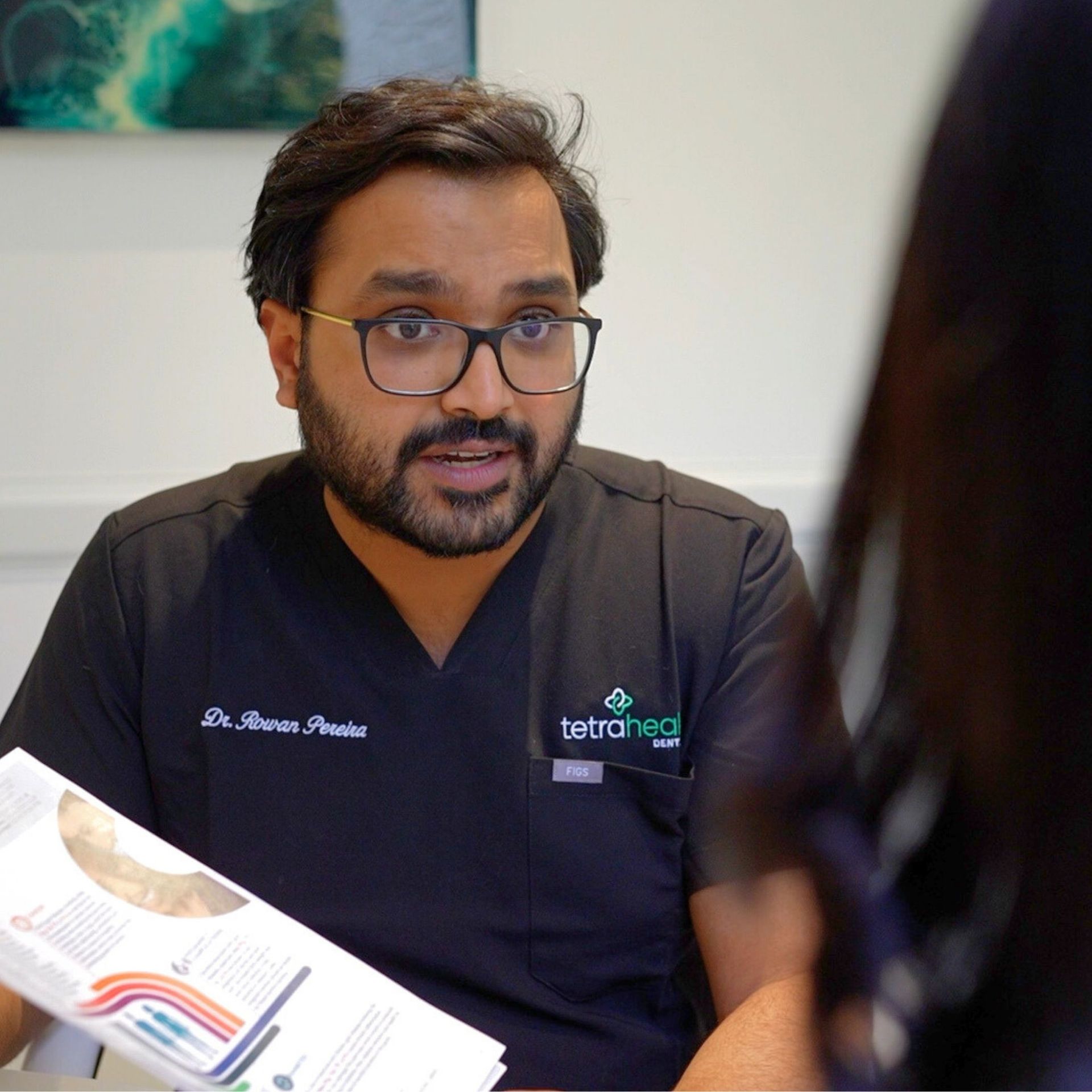 A doctor with glasses discusses a document with a patient. He is wearing a black top with
