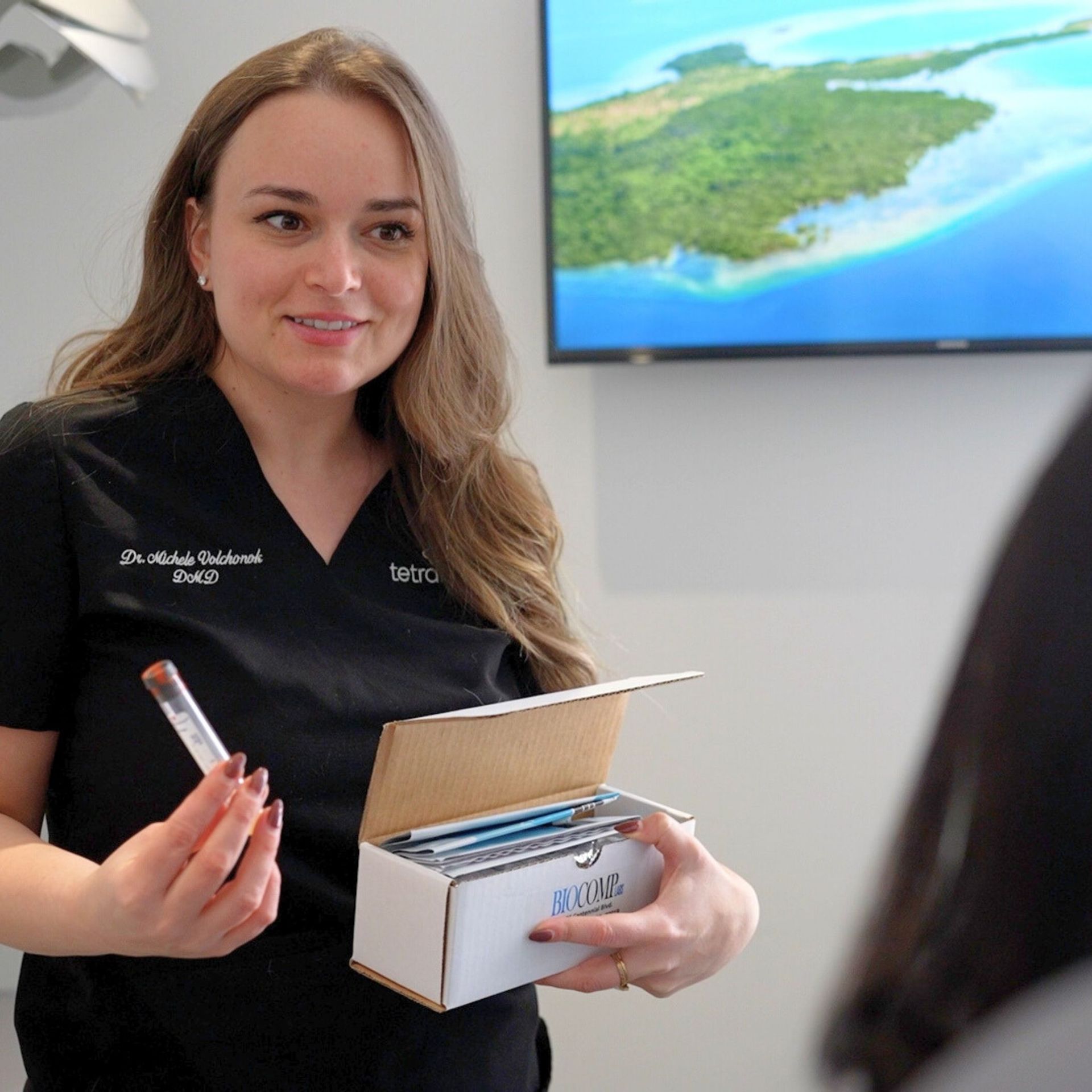 A woman in a black uniform holds a test tube and a box of medical supplies, likely explaining to a patient.