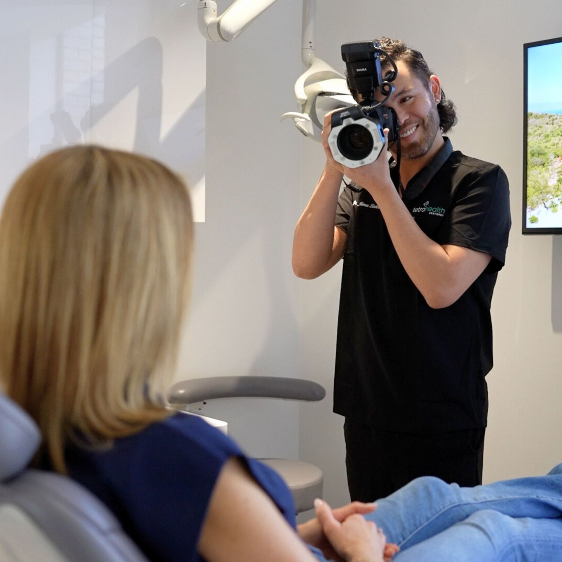 A dentist takes a photo of a patient in a dental chair. He smiles while holding a camera; the setting is a bright, modern office.