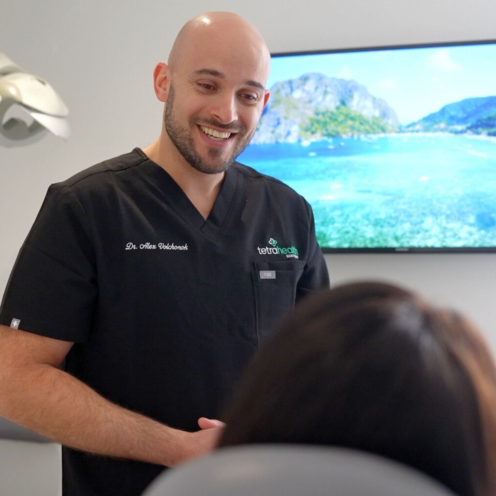 Male dentist in black scrubs smiling, interacting with a patient in a dental office, a tropical scene on the screen behind.