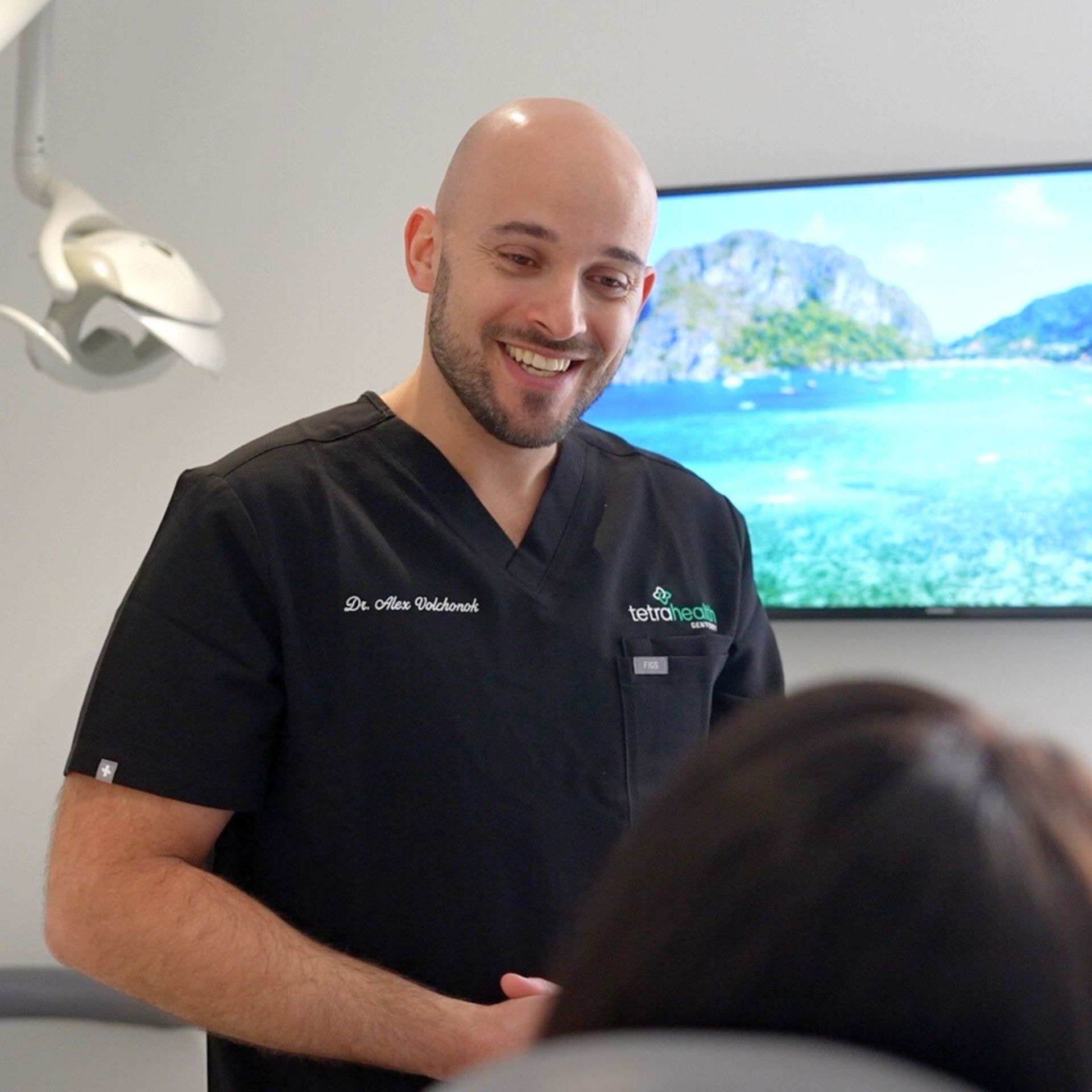 A dentist in black scrubs smiles, addressing a patient in a modern office. A screen with a tropical scene is visible.