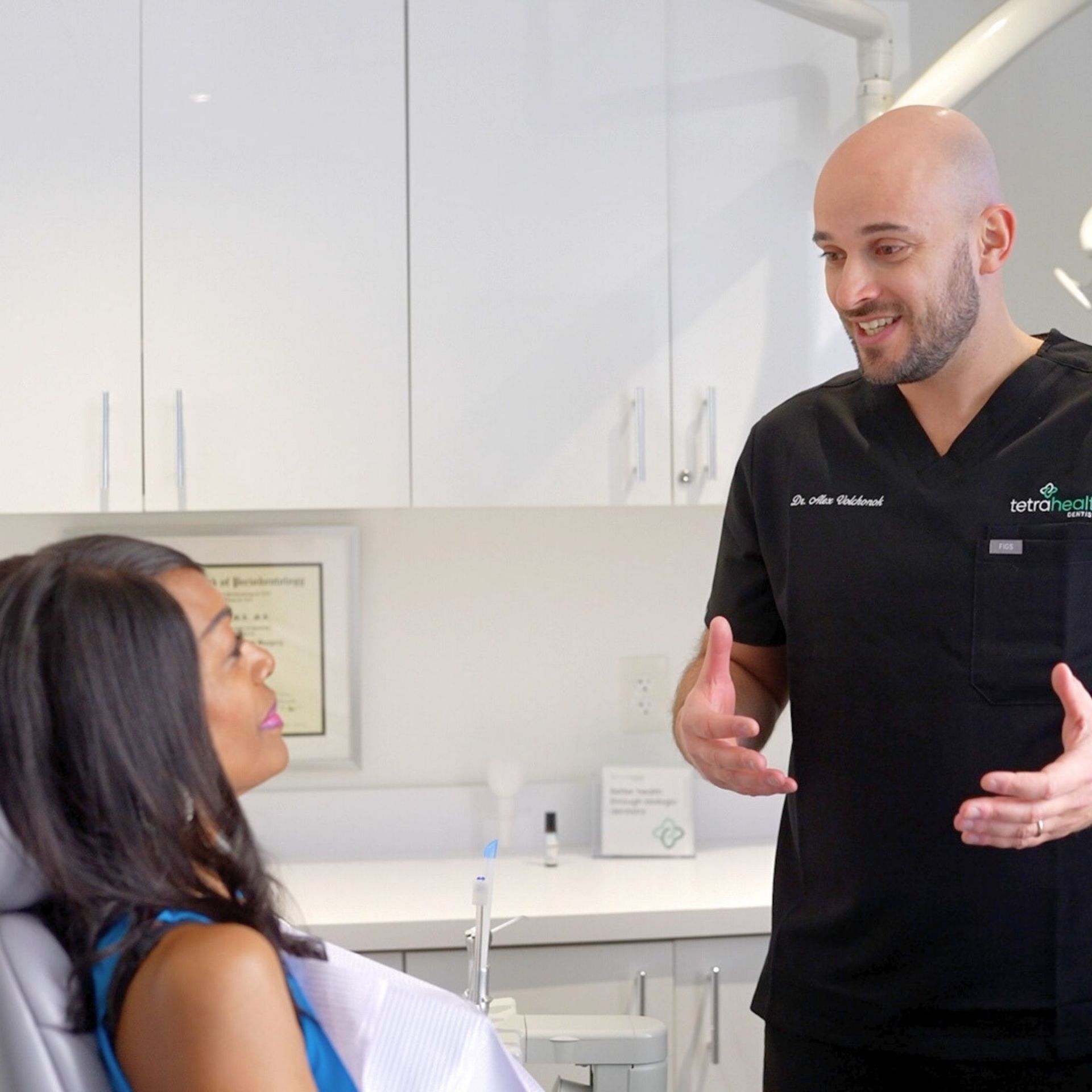 A dentist in black scrubs gestures as he talks to a woman in a dental chair; they are in a modern office.