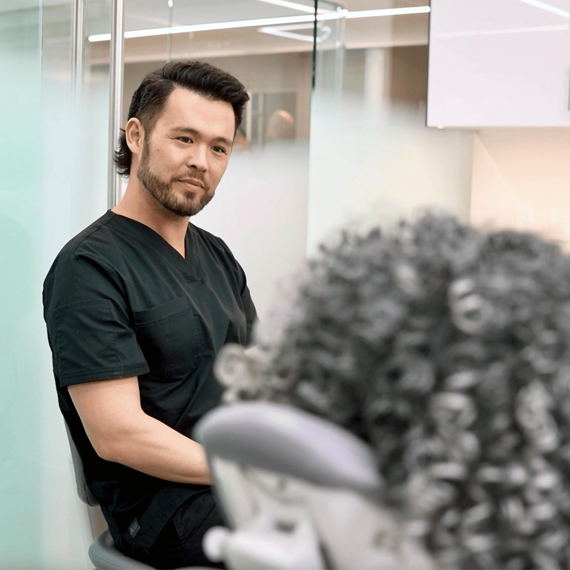 A male dentist in black scrubs leans towards a patient with curly hair in a dental chair. He appears attentive and slightly smiling.