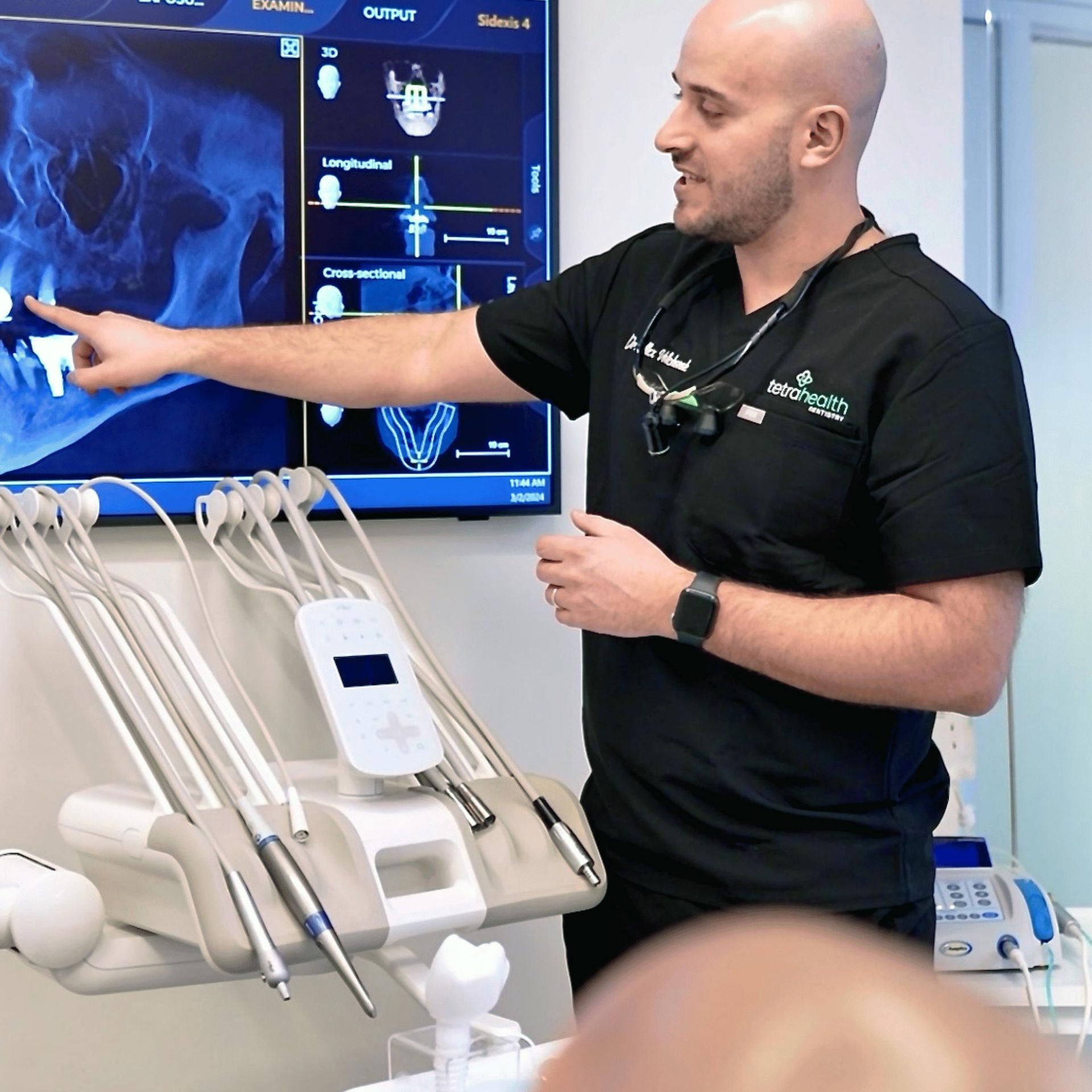 A bald dentist in a black uniform points to a dental X-ray on a monitor, explaining it to a patient in a modern office.