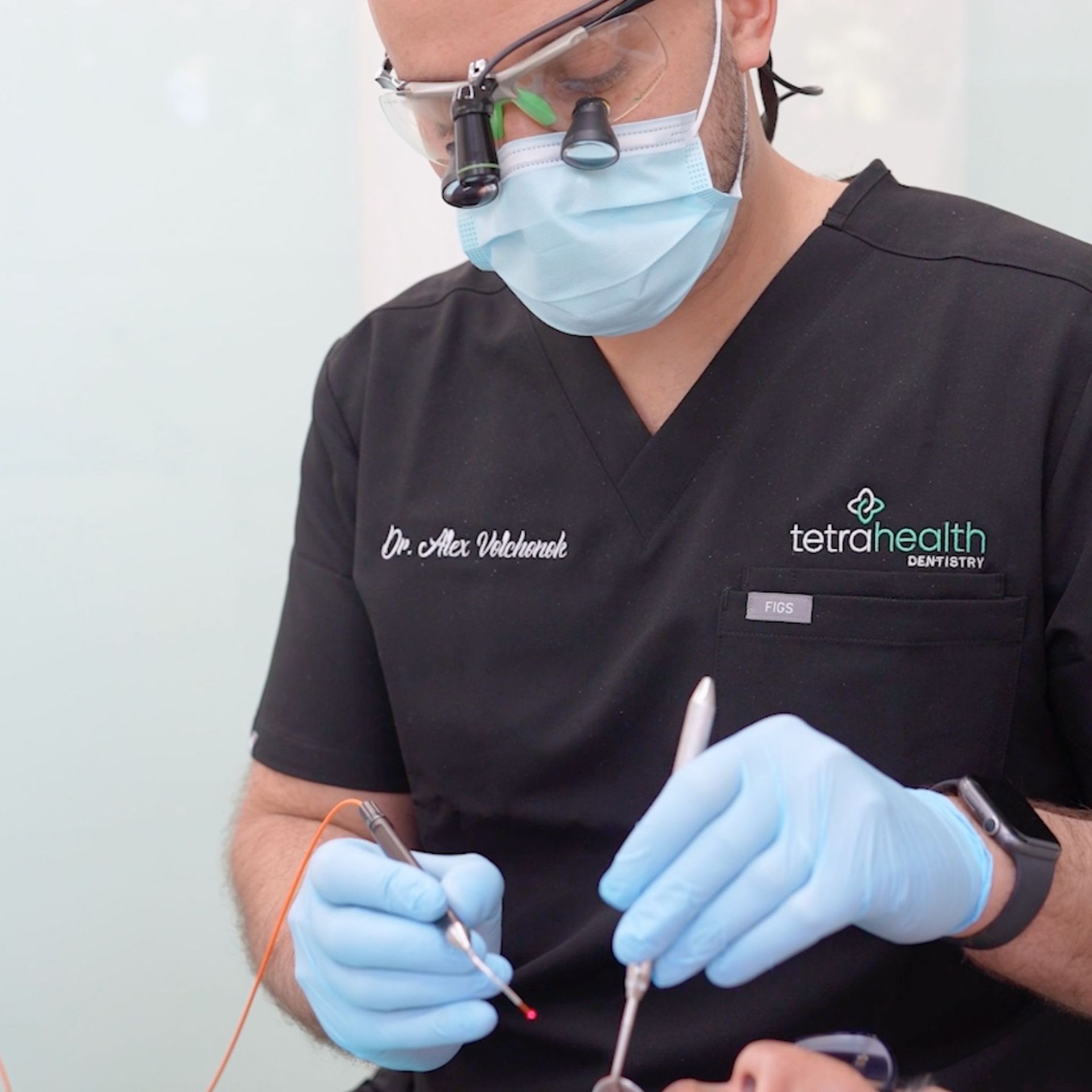 A dentist in scrubs and a mask using dental tools on a patient; the setting is a dental office.
