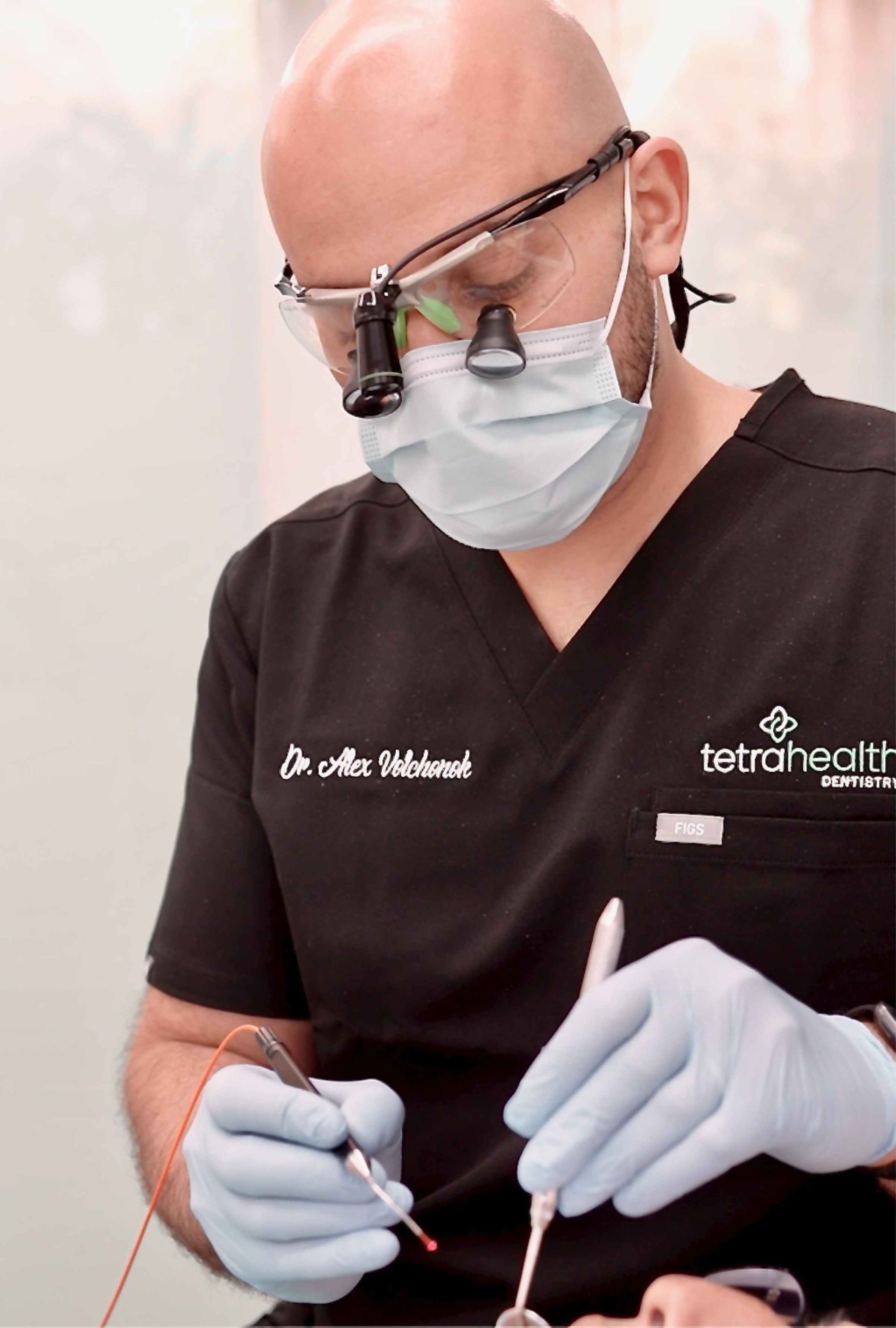 Dentist wearing a mask and loupes working on a patient's mouth. He holds a tool with a red laser pointer.