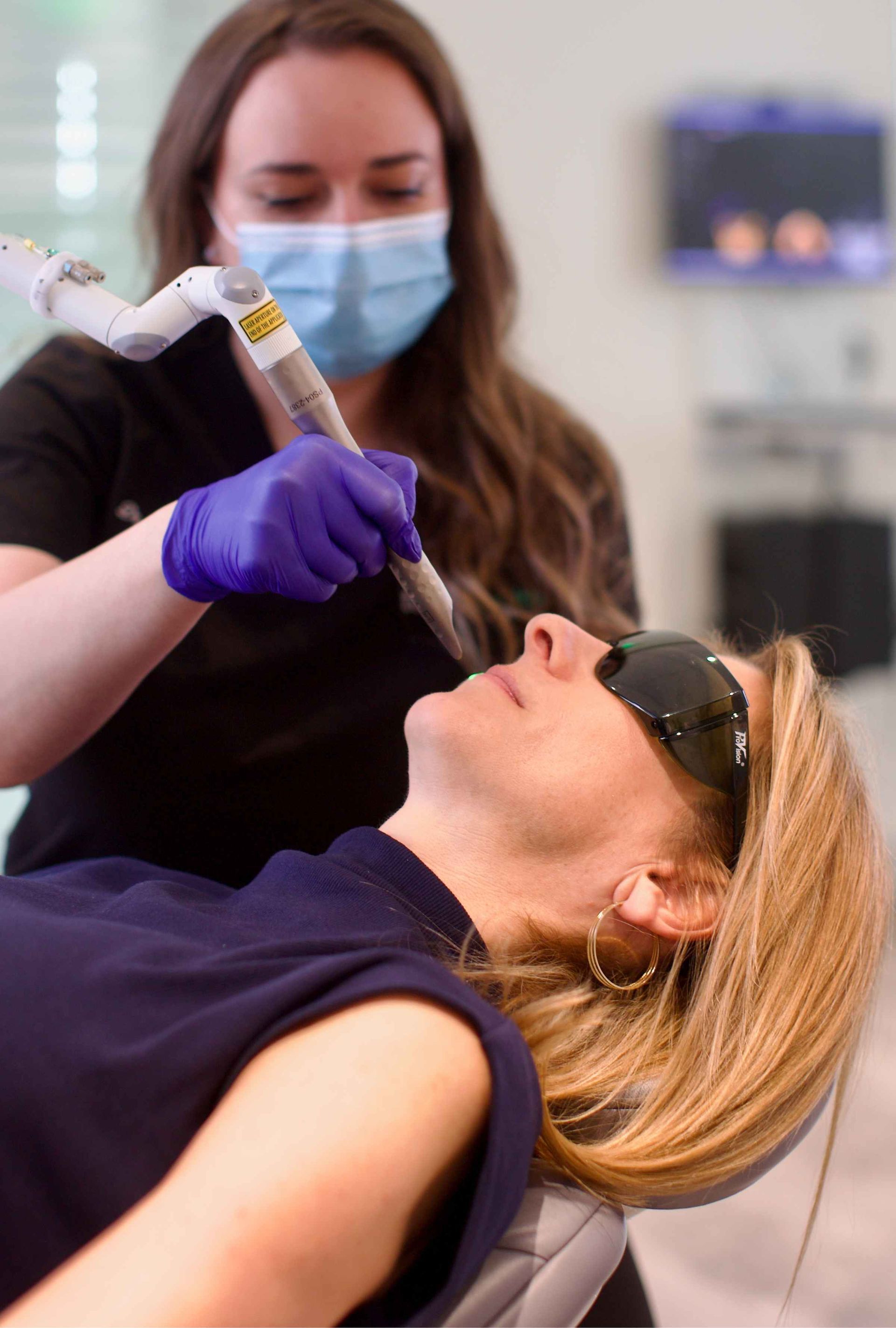 A medical professional in a mask and gloves uses a laser on a woman's face in a clinic setting. The patient wears protective eyewear.