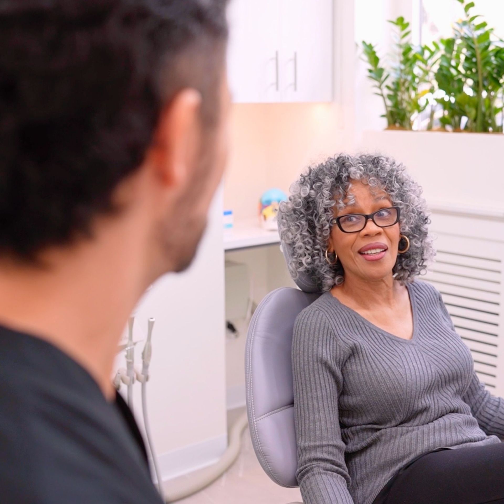 A dentist talking to an older woman patient in a light-filled office. The woman is seated, wearing glasses and a gray sweater.