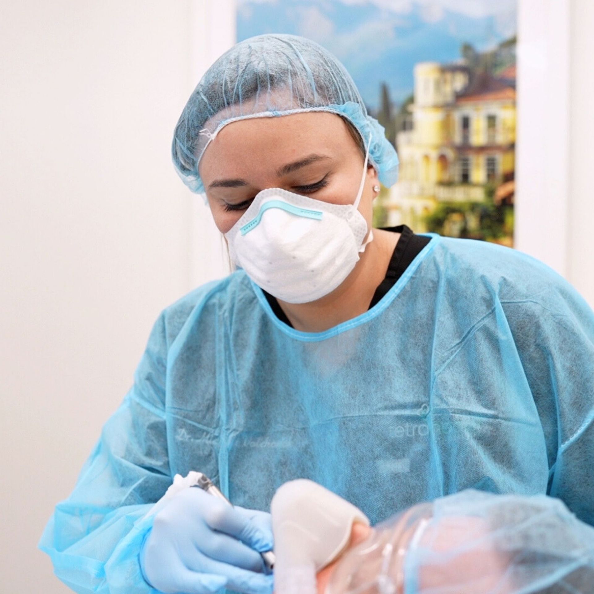 A person wearing a surgical mask, cap, gown, and gloves is performing a procedure, likely medical or cosmetic, in a clinic.