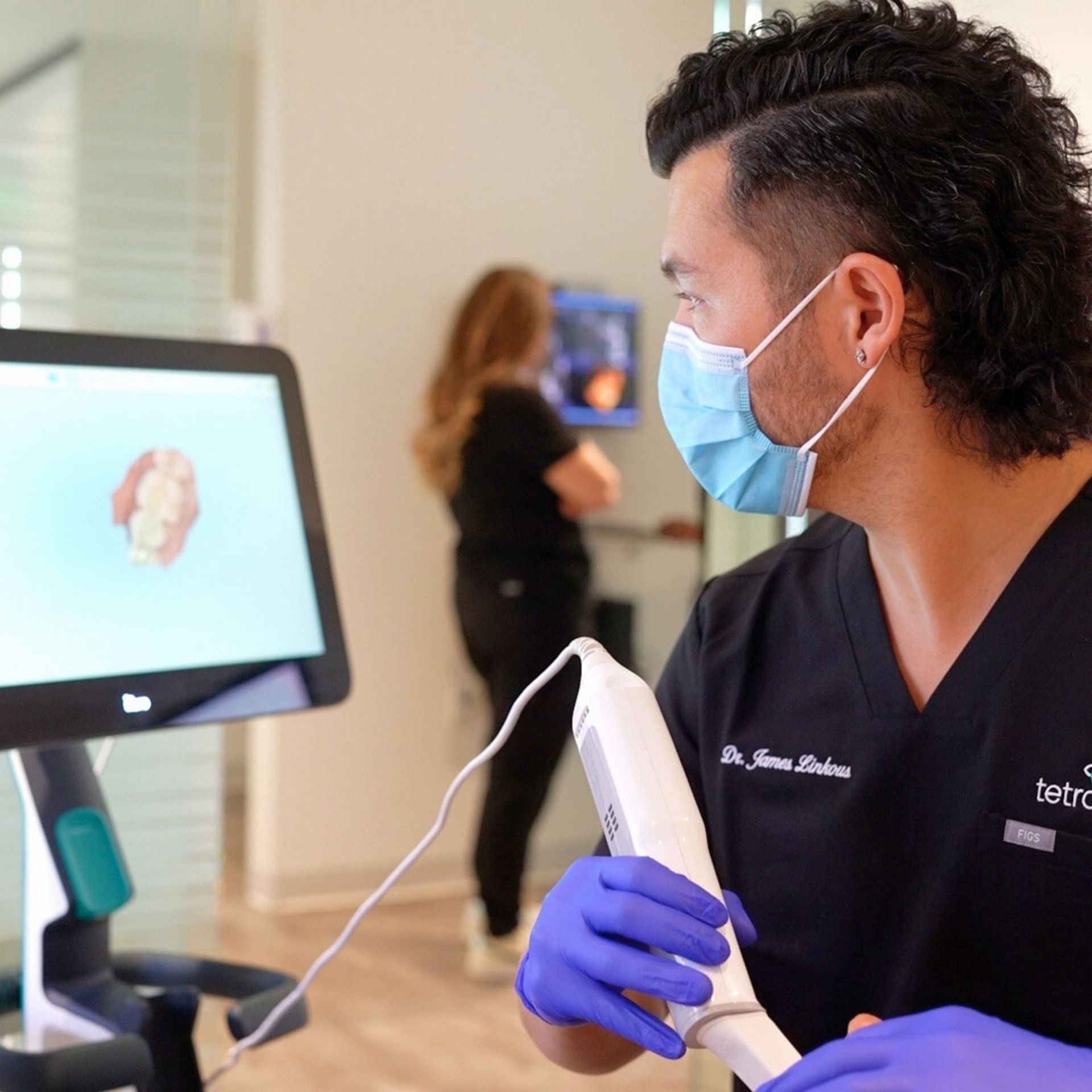 Dentist in blue gloves and mask examines a digital dental scan on a monitor, with a patient in the background.