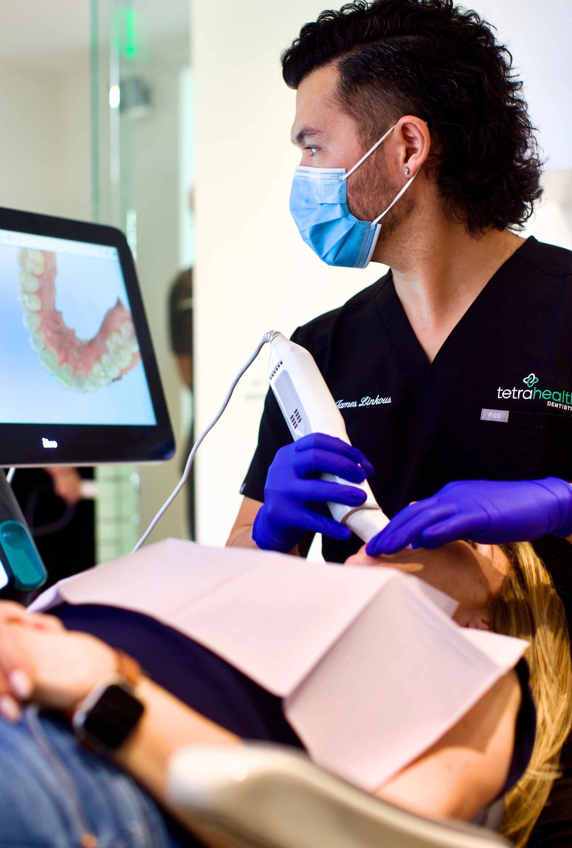 Dentist in black scrubs and mask using scanning device on patient, showing teeth scan on monitor.