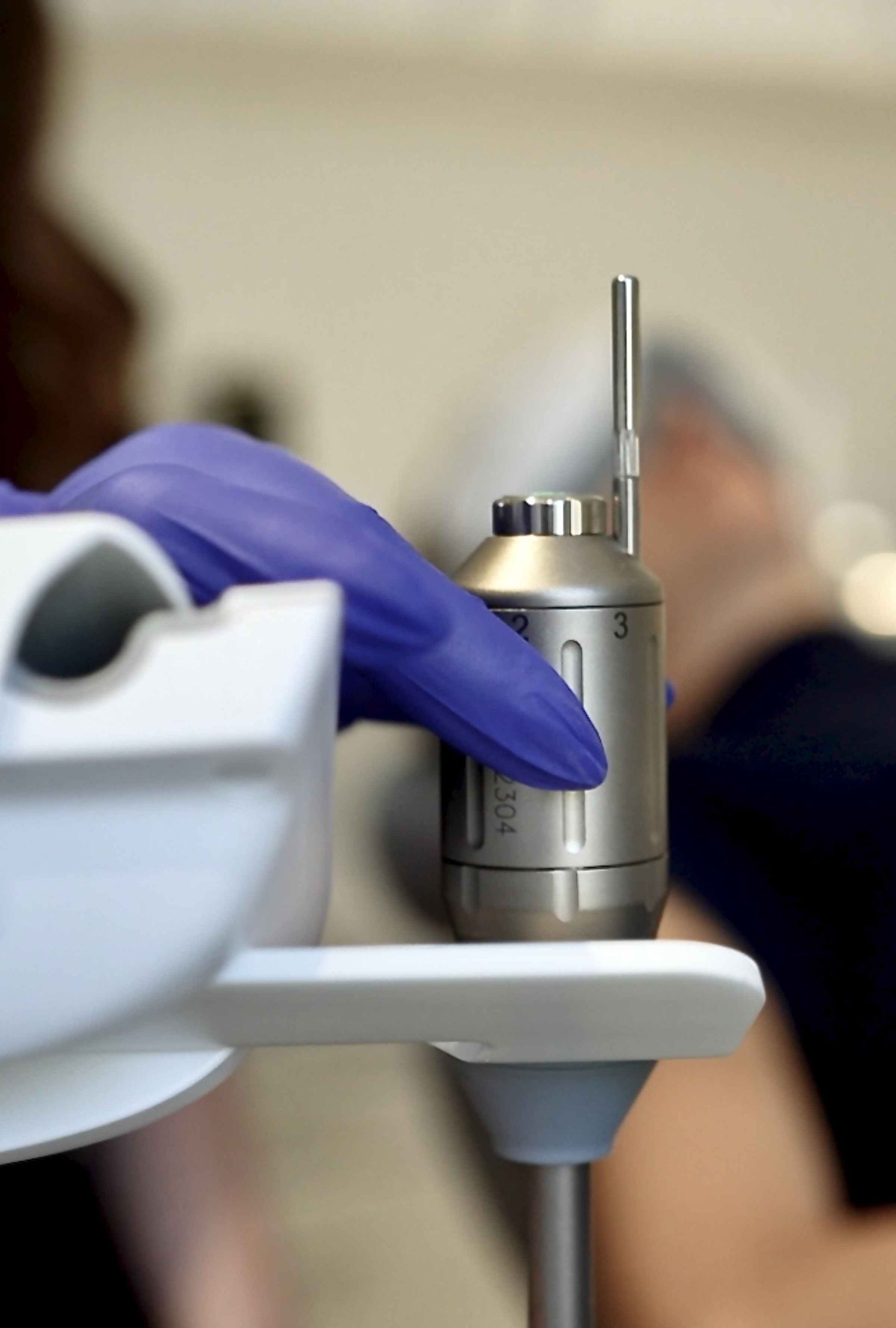Gloved hand adjusting a medical device. The device is metallic, with numbered settings; a person in the background.