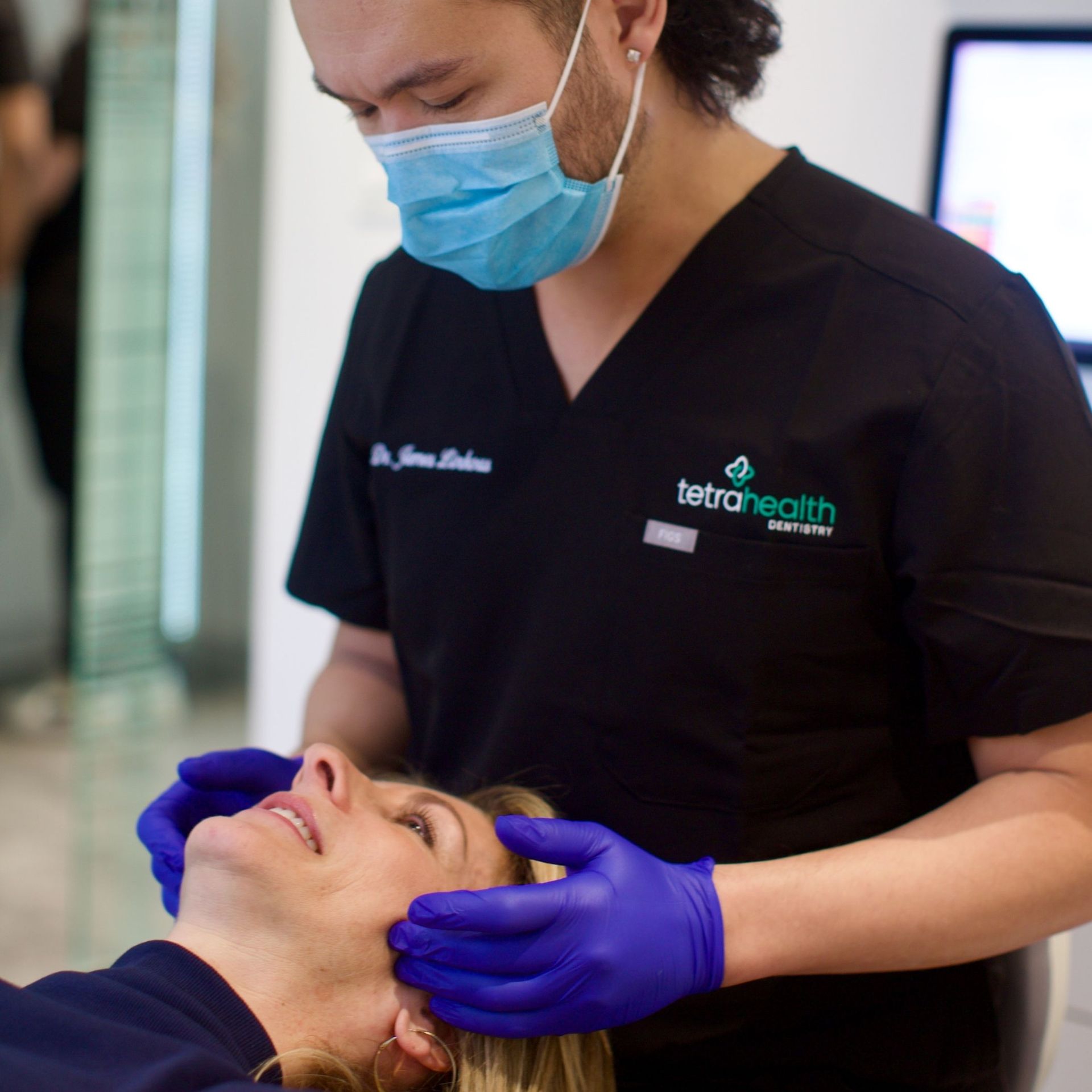 A healthcare provider in scrubs and a mask examining a woman's face with gloved hands; the woman reclines in a clinical setting.
