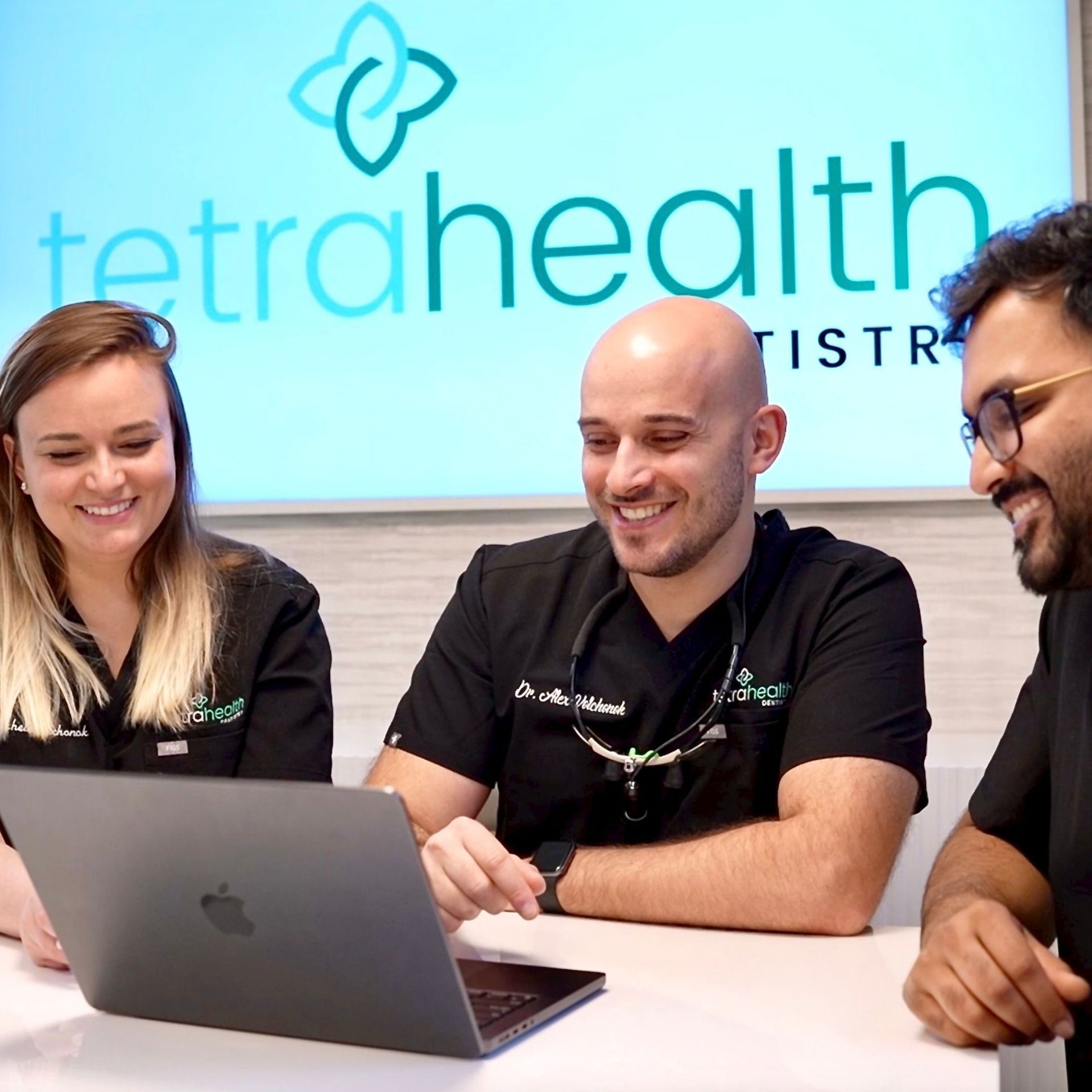 Three dental professionals at Tetra Health smile while looking at a laptop in an office. 