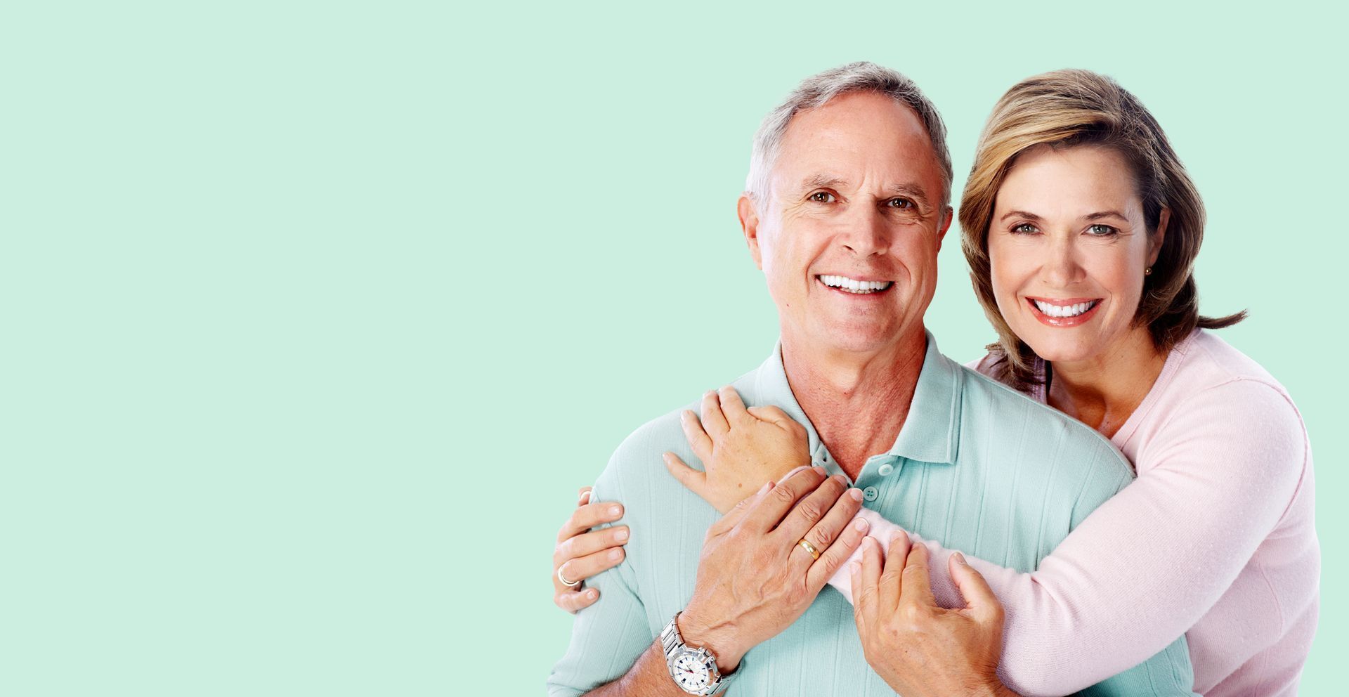 Smiling older couple embracing, woman behind the man with her arms around him, light green background.