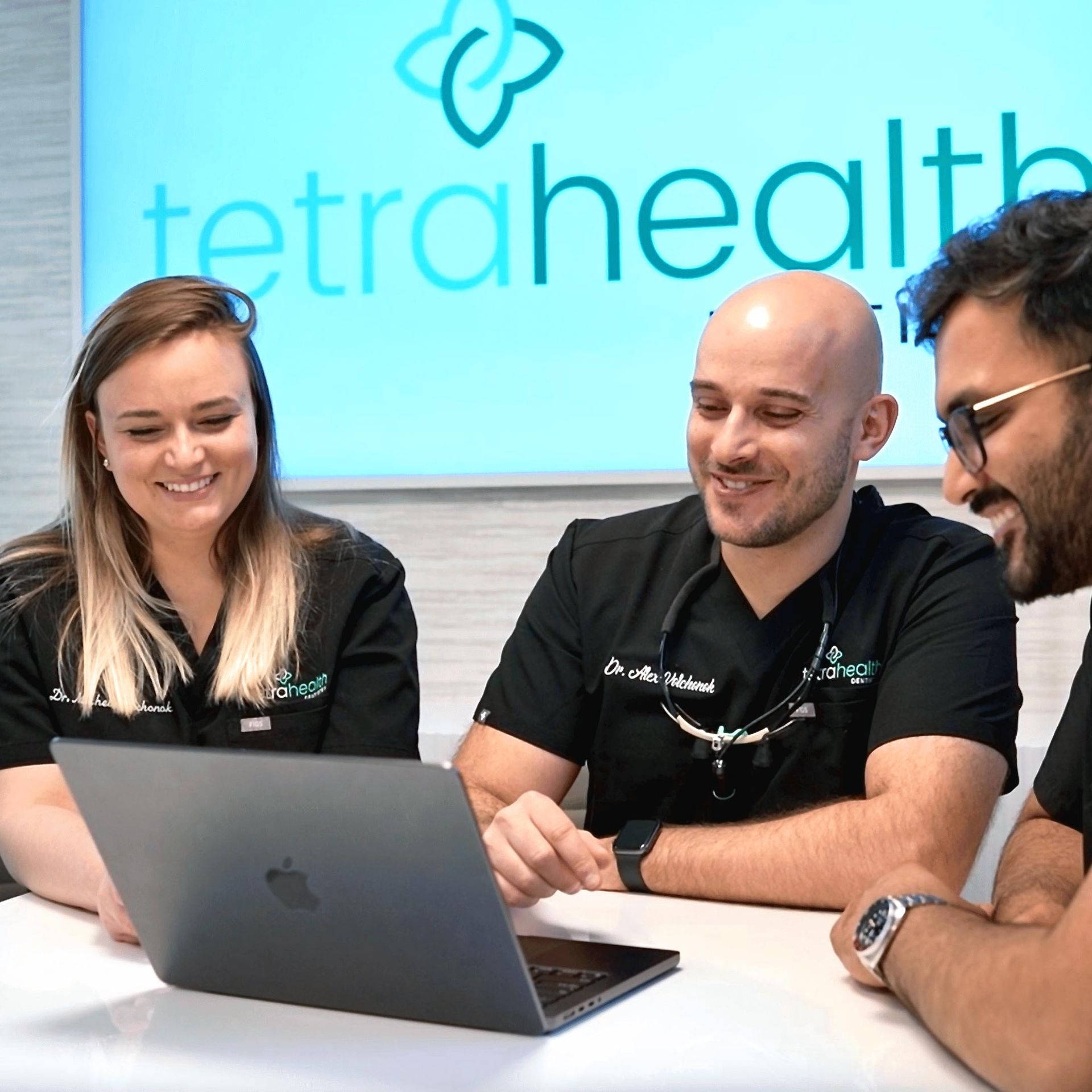 Three healthcare workers smiling and looking at a laptop, discussing. The Tetra Health logo is in the background.