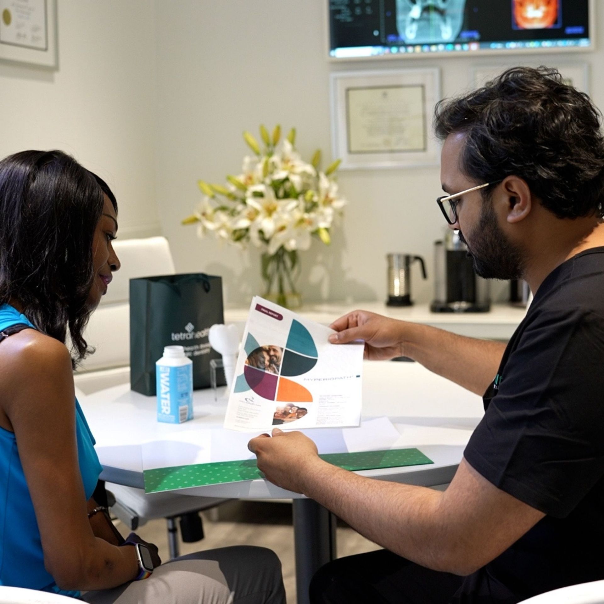 A doctor reviews paperwork with a female patient at a table in a clinic. The doctor is wearing glasses and scrubs.