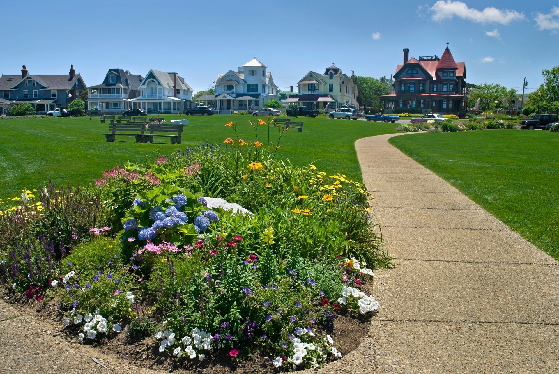 Park with flower bed, path, and houses. Green grass and blue sky.