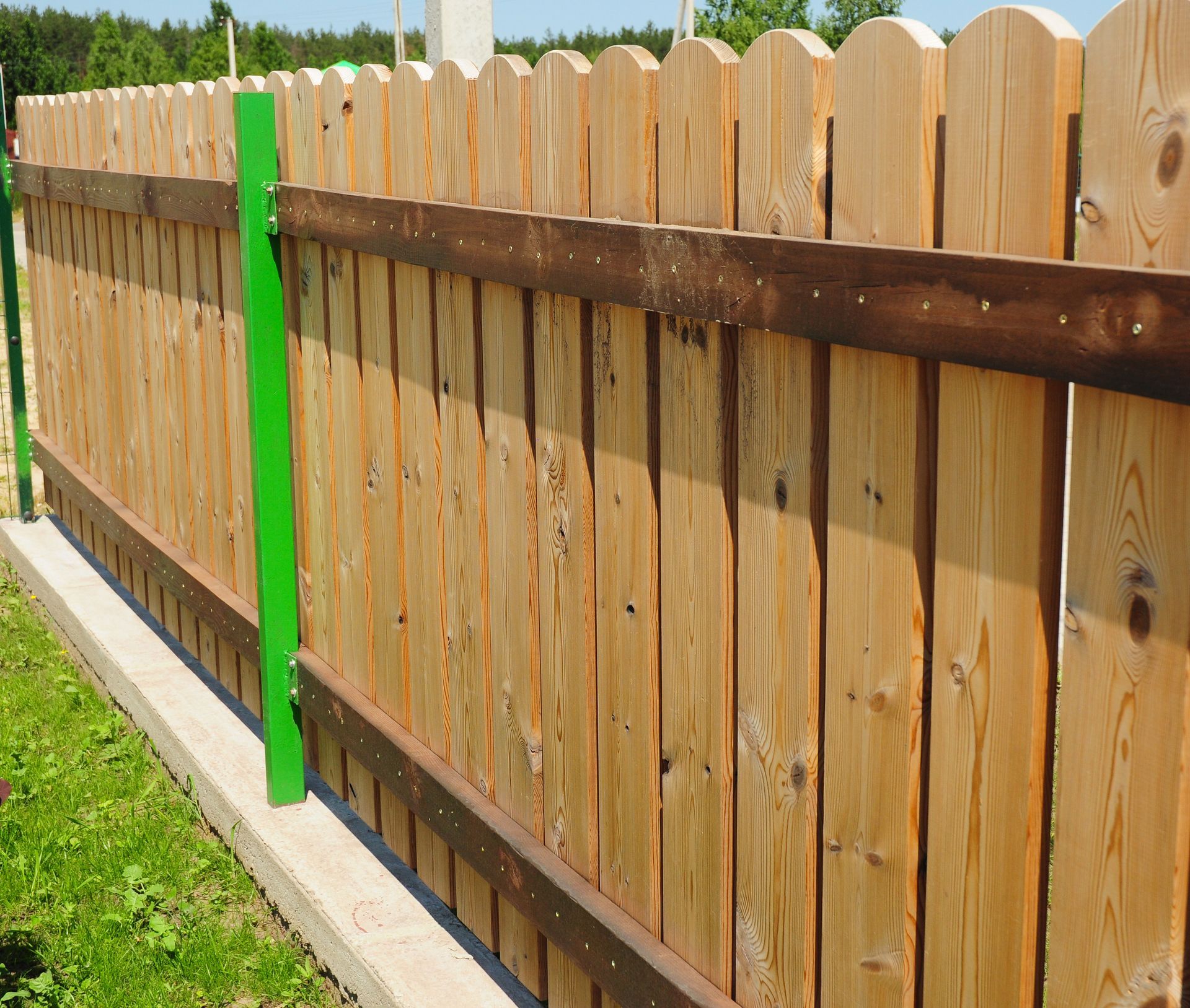 Wooden picket fence with scalloped tops, stained brown horizontal boards, and green support post.